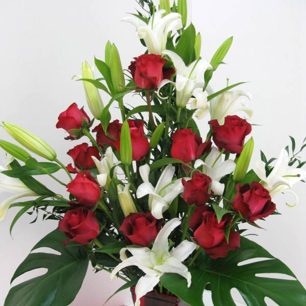 Tall arrangement of red roses and white lilies with large tropical leaves in a red glass vase.
