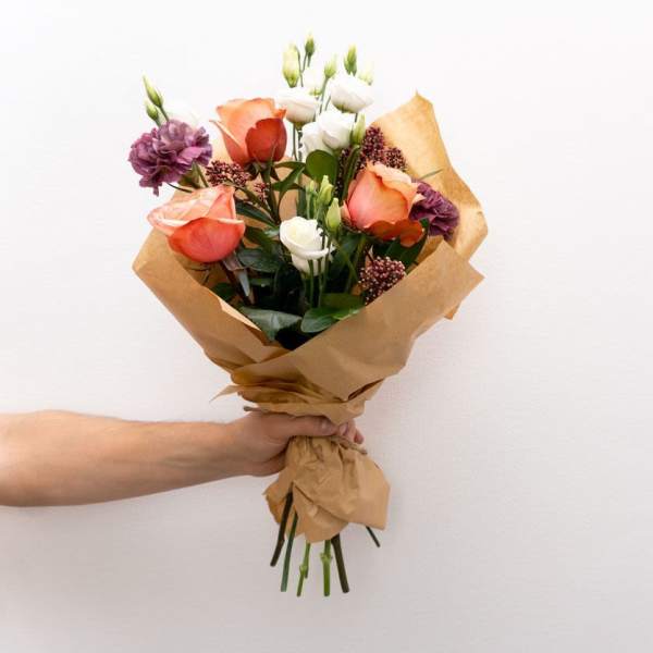Handheld bouquet of peach, white, and purple flowers wrapped in brown paper