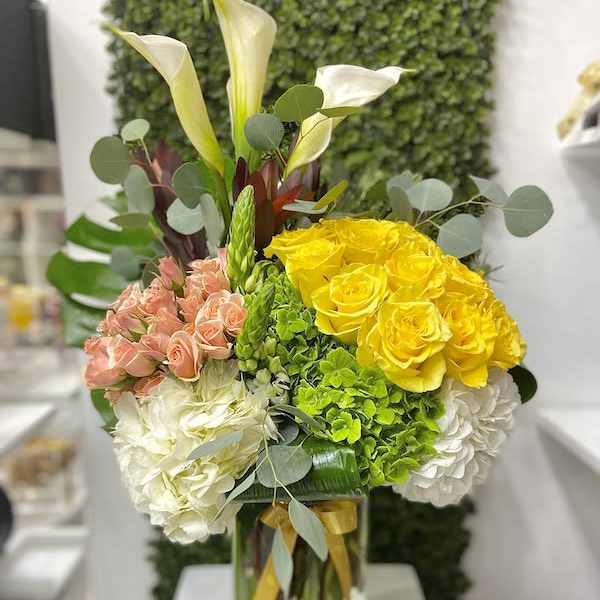 Tall arrangement of yellow roses, peach spray roses, white hydrangeas, and calla lilies in a glass vase
