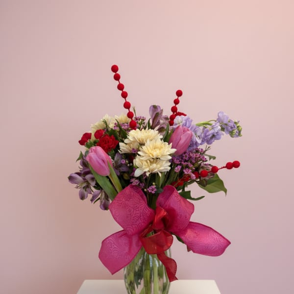 Mixed bouquet of pink, purple, and cream flowers in a glass vase with a large pink bow