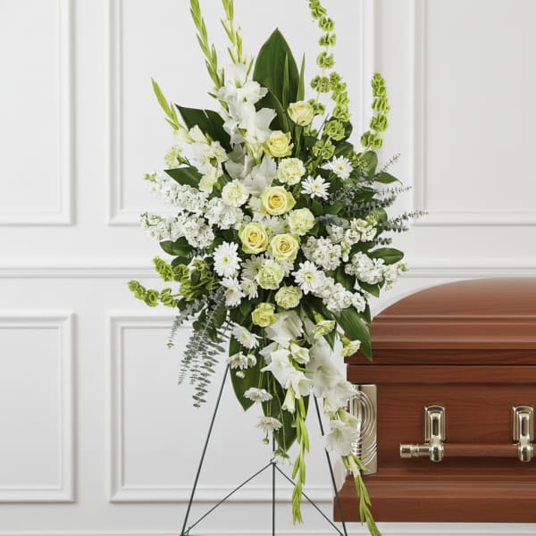 Tall white and pale yellow standing floral spray on an easel beside a wood casket