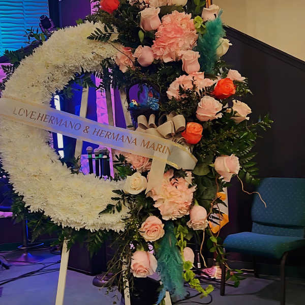 Large crescent-shaped white standing wreath with pink and orange roses on an easel indoors