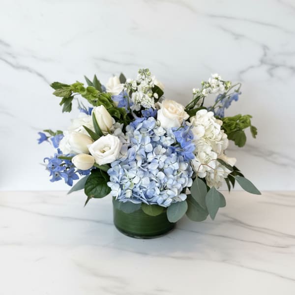 Low arrangement of white roses, tulips, and blue hydrangeas in a green glass cylinder vase