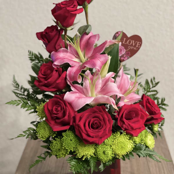 Red roses and pink lilies arranged in a red vase with a heart tag