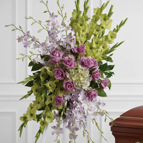 Standing spray of green gladiolus, lavender roses, and white orchids on a white easel beside a casket.