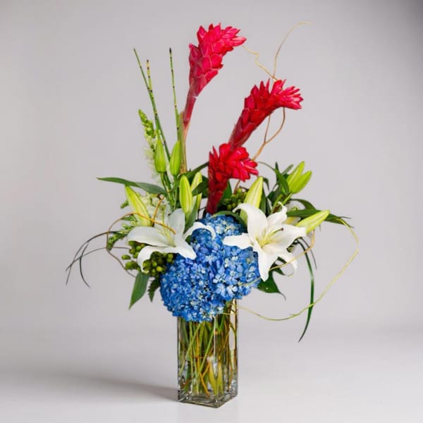 Tall vase arrangement with blue hydrangea, white lilies, and red tropical blooms