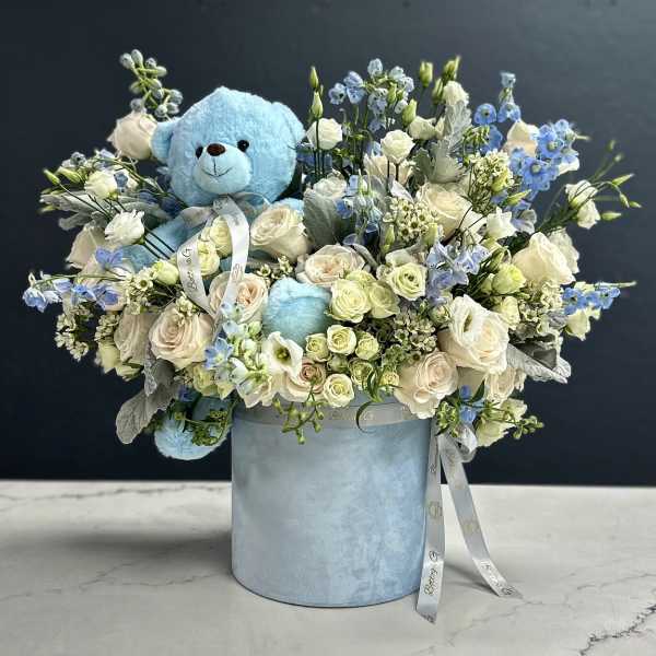 Blue teddy bear in a pastel flower arrangement with white and blue blooms