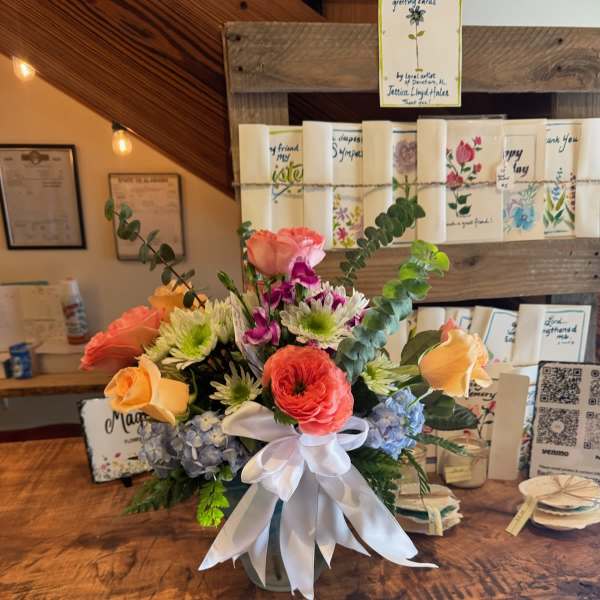 Mixed bouquet of roses, ranunculus, hydrangea, and daisies in a vase with a white ribbon