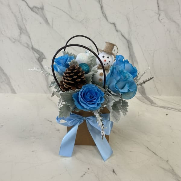 Blue roses in a small gift basket with a snowman ornament and pinecone