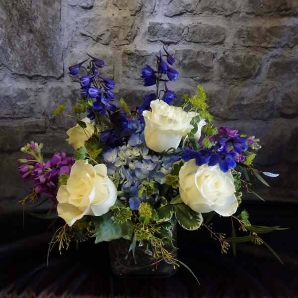 Bouquet of white roses, blue flowers, and purple blooms in a glass vase