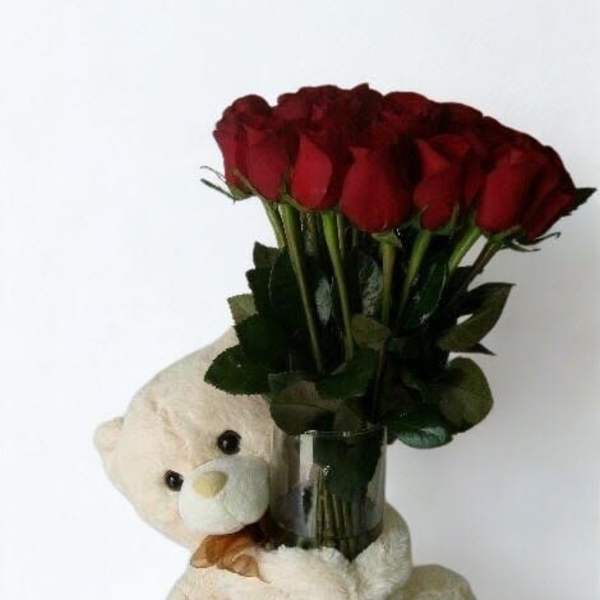 Red roses in a glass vase with a teddy bear hugging it