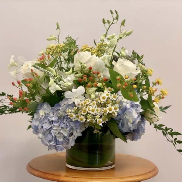 Low glass vase of white roses, blue hydrangeas, and small white daisies with berries on a wooden table