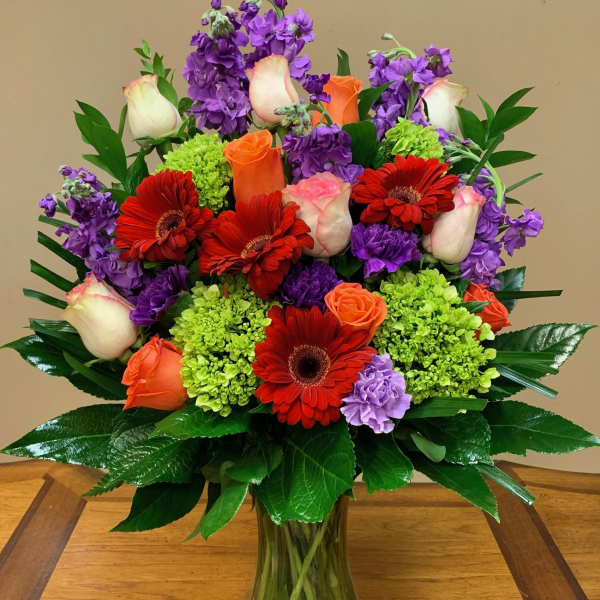 Mixed bouquet of red gerbera daisies, orange and white roses, and purple flowers in a glass vase