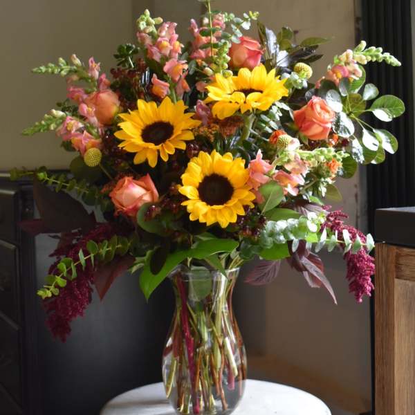 Mixed bouquet with sunflowers, peach roses, and pink snapdragons in a clear glass vase