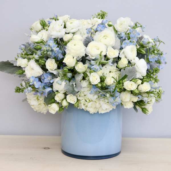 Round arrangement of white and light blue flowers in a light blue cylinder vase