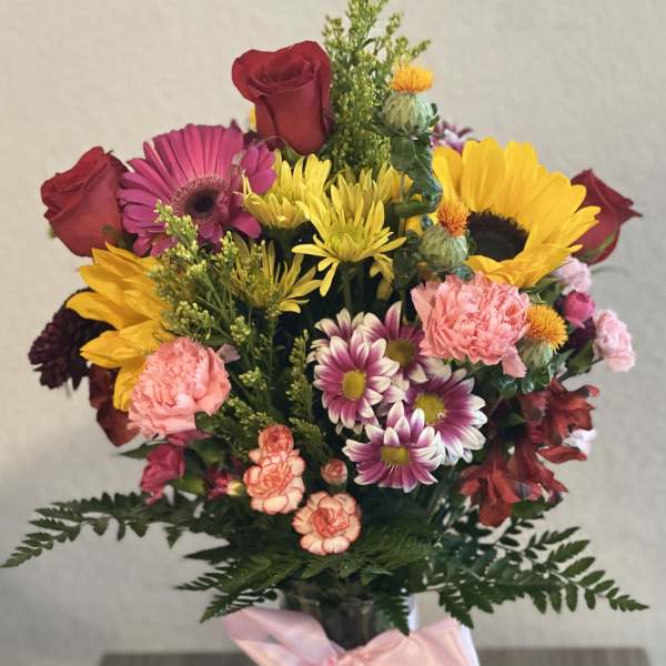 Mixed bouquet of roses, sunflowers, daisies, and carnations in a glass vase with a pink ribbon.