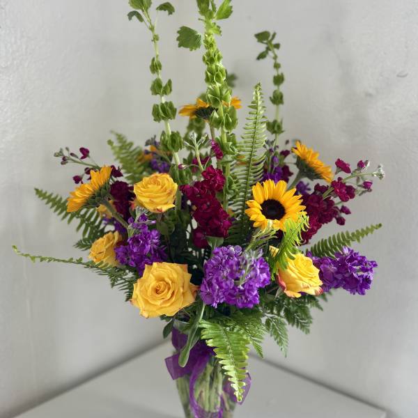 Mixed bouquet of yellow roses, sunflowers, and purple flowers in a glass vase
