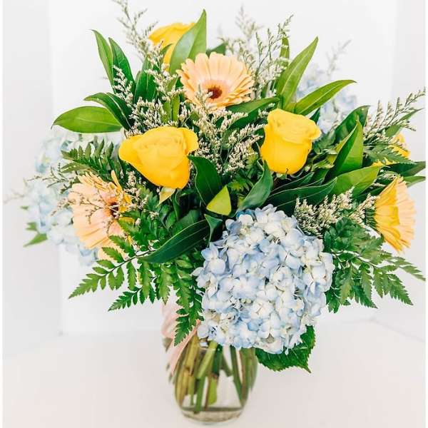 Bouquet of yellow roses, gerbera daisies, and blue hydrangea in a glass vase