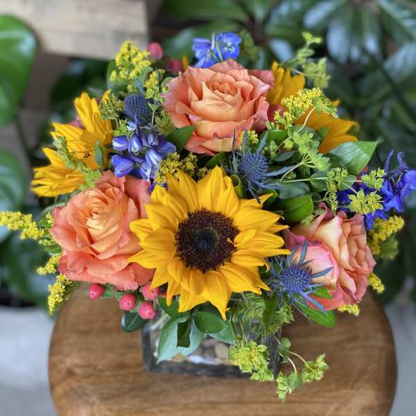 Bouquet of sunflowers, peach roses, and blue flowers in a glass vase
