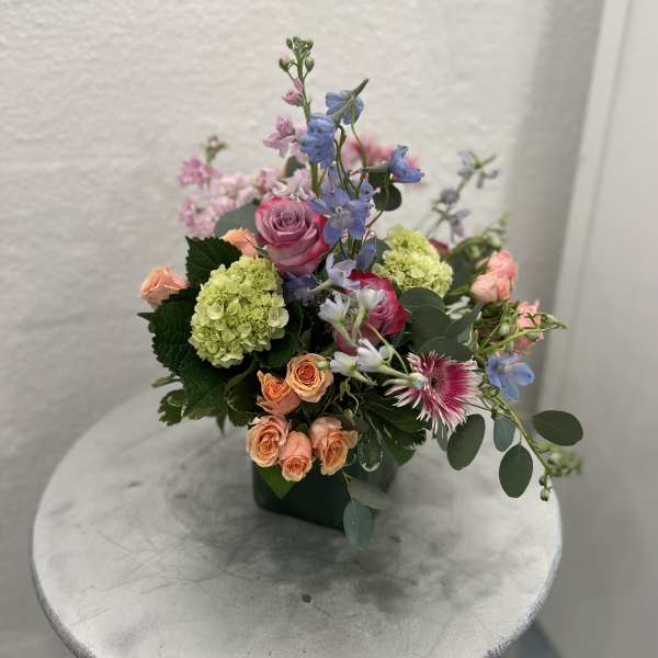 Pastel arrangement of pink and peach roses, green hydrangea and blue blooms in a green square vase