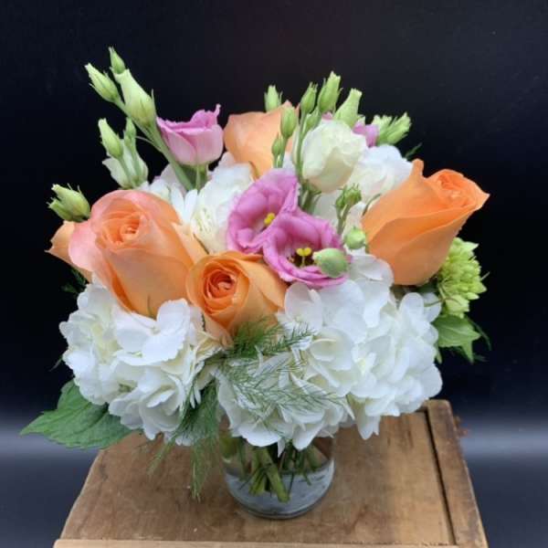 Short arrangement of peach roses, white hydrangeas, and pink lisianthus in a clear glass vase.