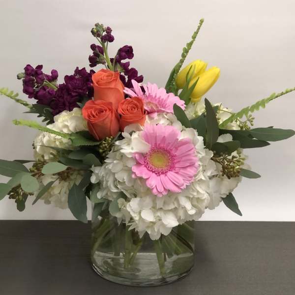 Low mixed arrangement of white hydrangeas, pink gerberas, orange roses, and yellow tulips in a glass vase