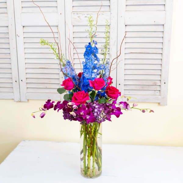 Tall vase arrangement with blue flowers, pink roses, and purple orchids