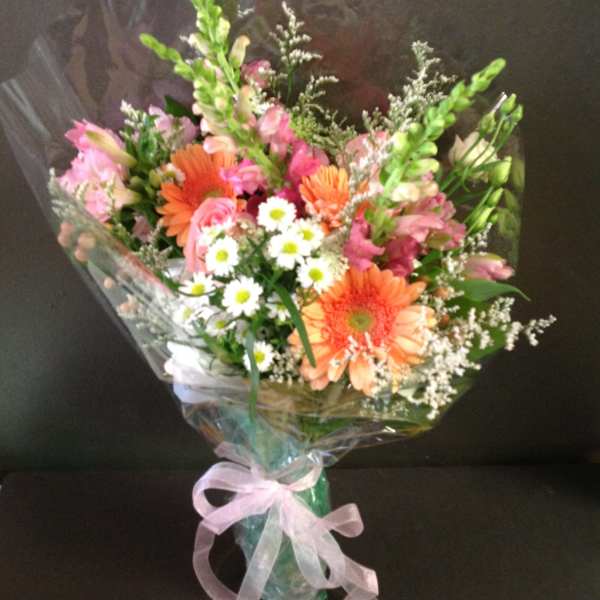 Hand-tied bouquet of orange gerbera, pink roses, and white daisies wrapped in cellophane with a pink bow