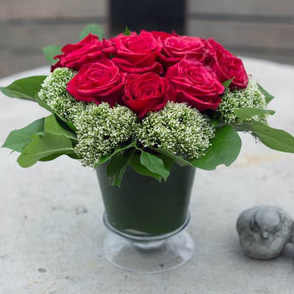 Red roses arranged in a dark vase with green foliage and white filler flowers