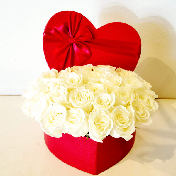 White roses arranged in a red heart-shaped box with a red ribbon bow
