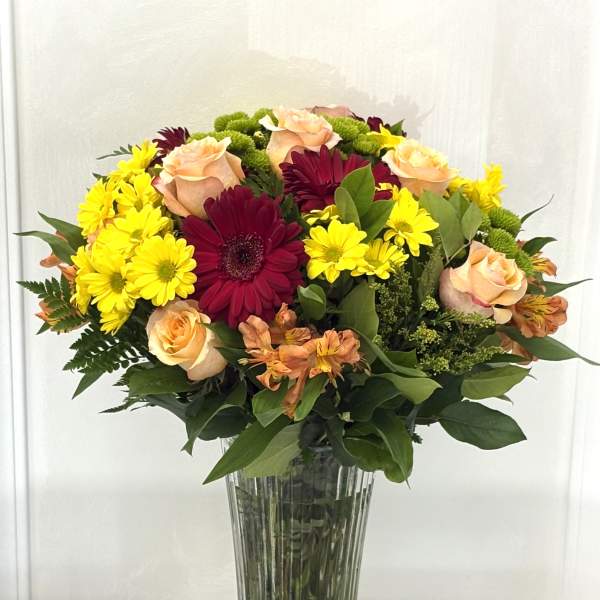 Mixed bouquet of yellow daisies, peach roses, and burgundy gerberas in a glass vase