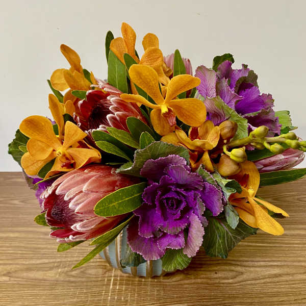 Mixed bouquet with orange orchids, purple kale, and red protea in a low container