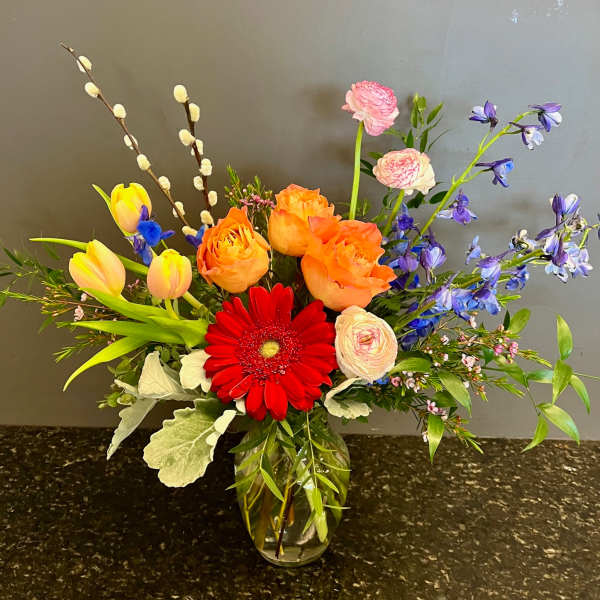 Mixed bouquet in a glass vase with red gerbera, orange roses, yellow tulips, and purple flowers