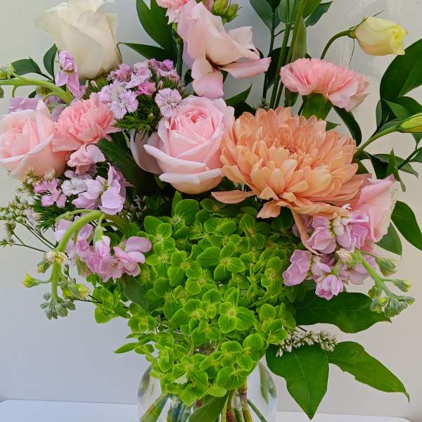 Tall glass vase of pink roses, carnations, lisianthus, peach mum, and green hydrangea.