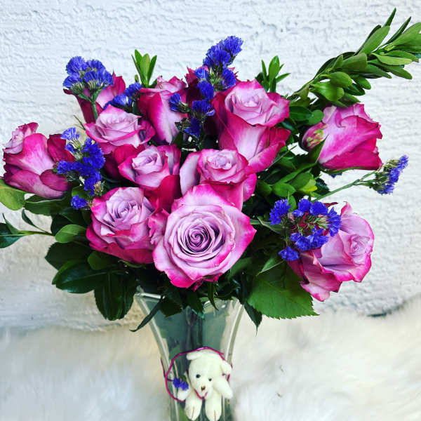 Pink roses and blue filler flowers in a clear glass vase with a small teddy bear
