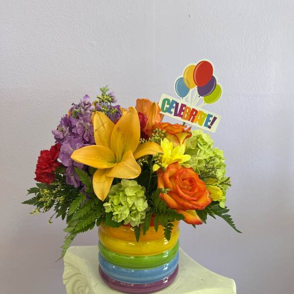 Colorful flower arrangement in a rainbow-striped vase with a "CELEBRA EL!" topper