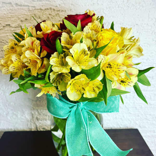 Yellow alstroemeria and red roses in a glass vase with a teal ribbon