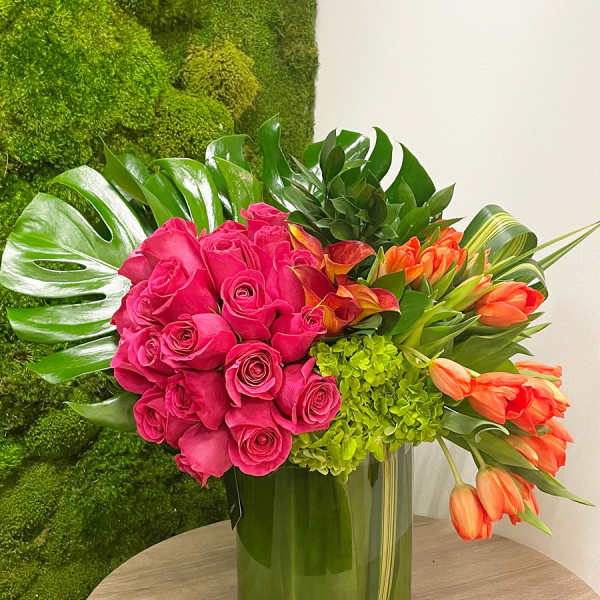 Bright pink roses, orange tulips, and green hydrangeas arranged with large tropical leaves in a clear glass cylinder vase