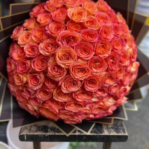Large spherical bouquet of orange and pink roses wrapped in dark fan-folded paper.