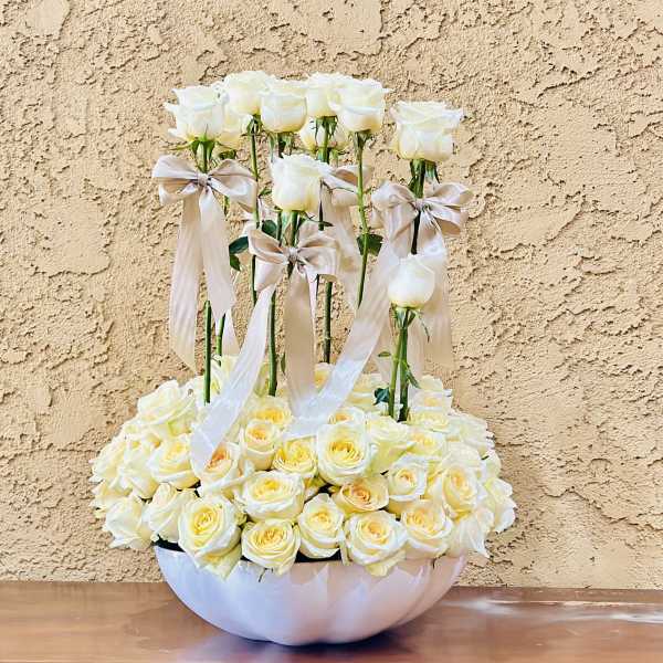 White roses arranged in a low bowl with tall rose stems and satin bows
