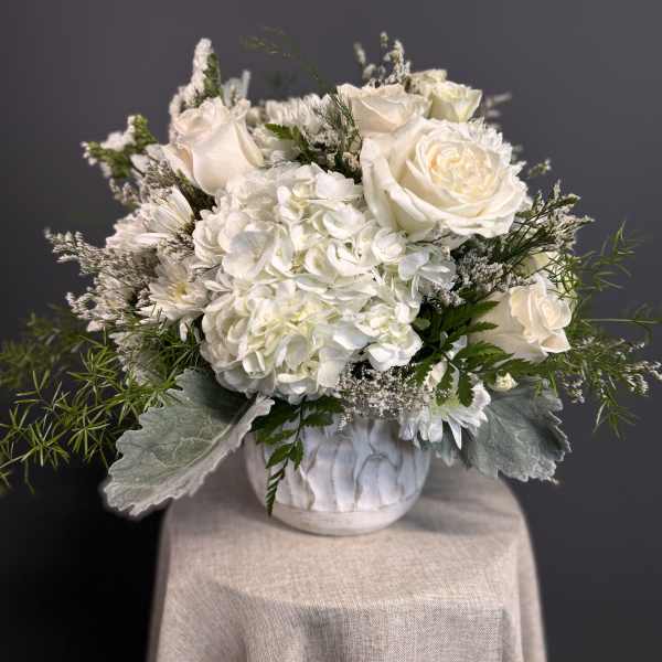 Low white arrangement of roses and hydrangeas in a white ceramic pot on a draped pedestal