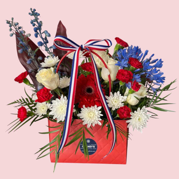Red, white, and blue floral arrangement in a red gift box with ribbon