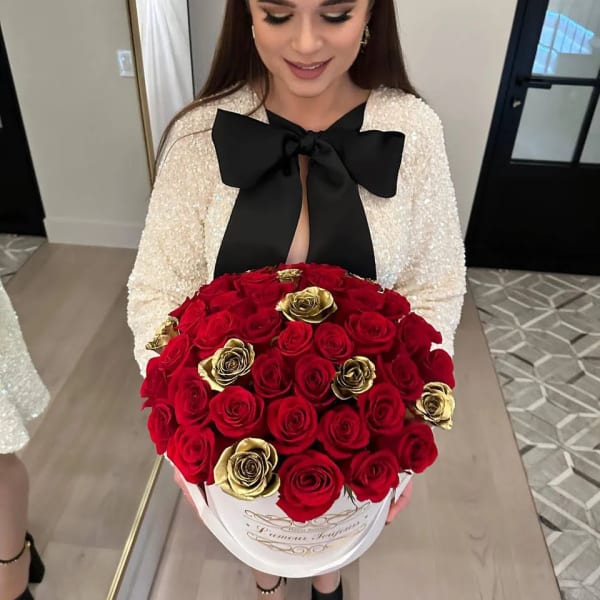 Round box of red and gold roses held in front of a woman in a dress