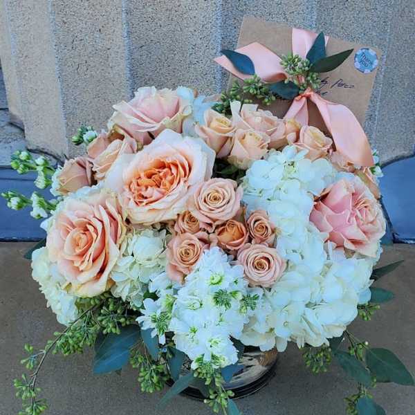 Low arrangement of peach roses and white hydrangeas in a glass vase with a card tied by a ribbon
