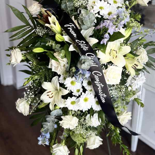 Tall standing spray of white lilies, roses, and daisies with light blue flowers and a black ribbon.