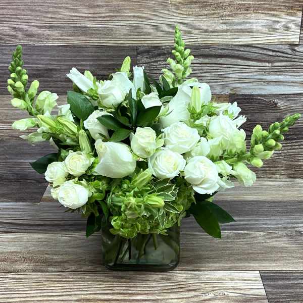 White roses and snapdragons with green accents arranged in a clear glass cube vase