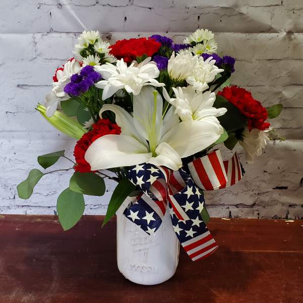Bouquet of white lilies, red carnations, and purple daisies in a white vase with patriotic ribbon