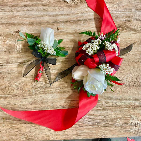 Two floral corsages with red ribbon and white roses on a wood surface