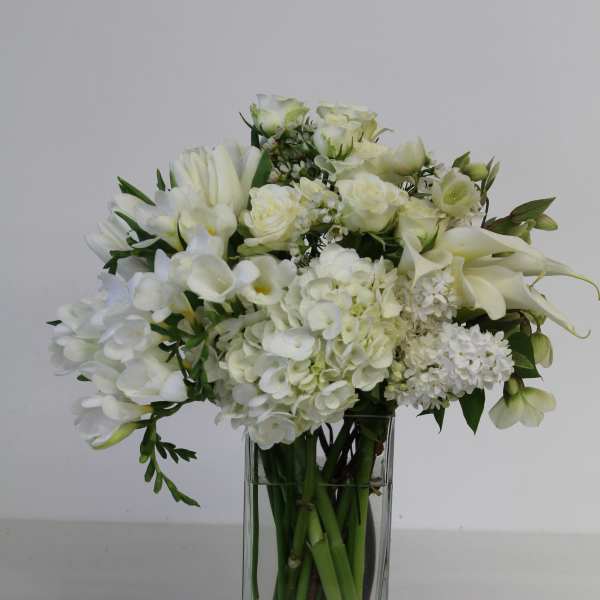 Tall arrangement of white flowers including roses, hydrangeas, and lilies in a clear glass vase