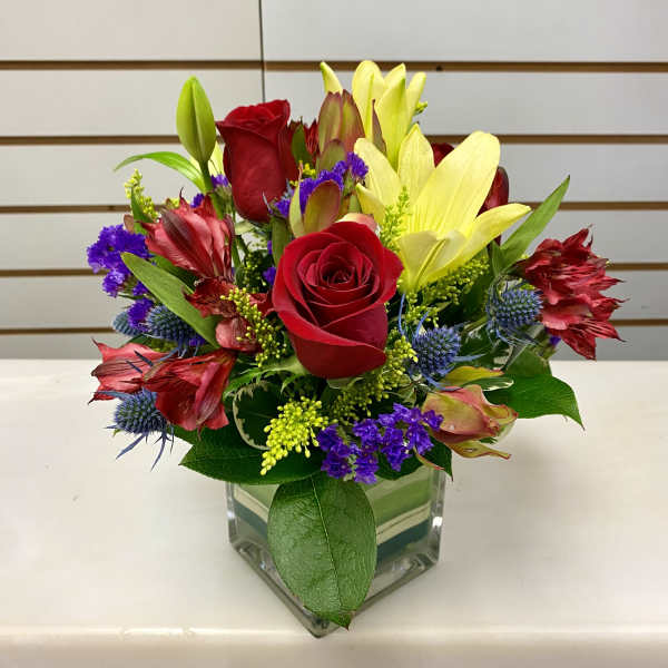 Compact arrangement of red roses, yellow lilies, and mixed accent flowers in a clear glass cube vase